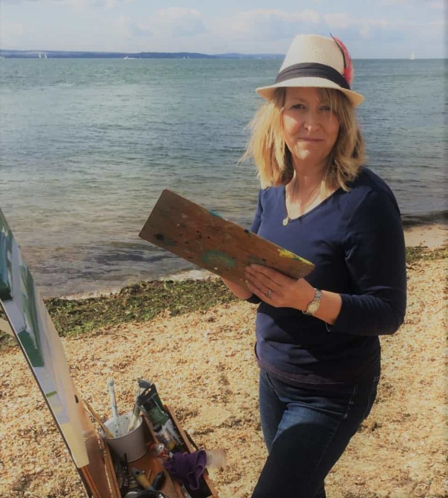 Artist Carolyn Tyrer standing by the sea and infront of her easel smiling and painting a picture